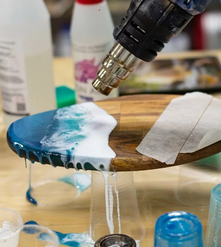 Wooden spoon with blue and white resin being applied using a stearnel hot air blower on a blurred background.